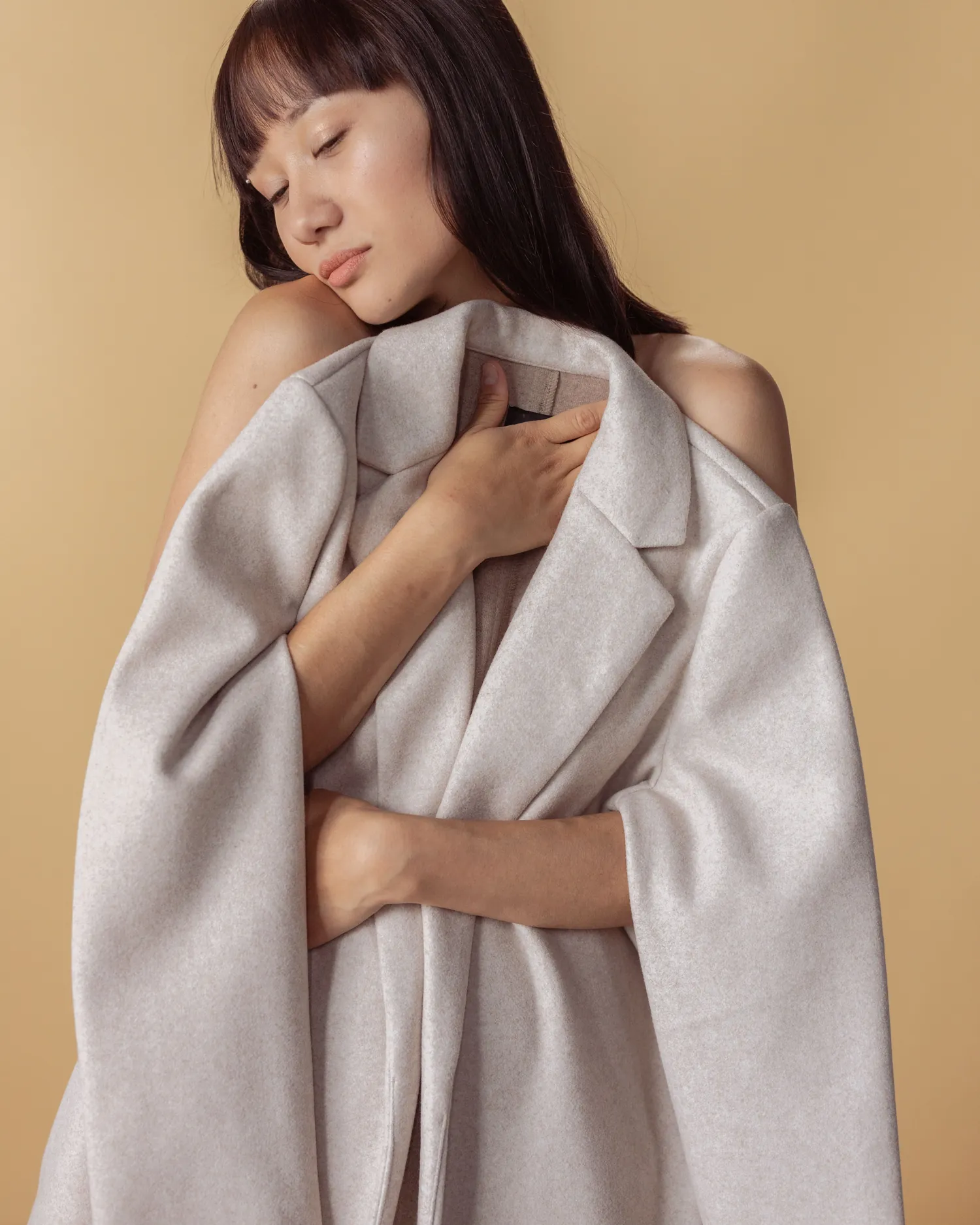Model holding an undyed lambswool coat closed against a warm ochre ground
